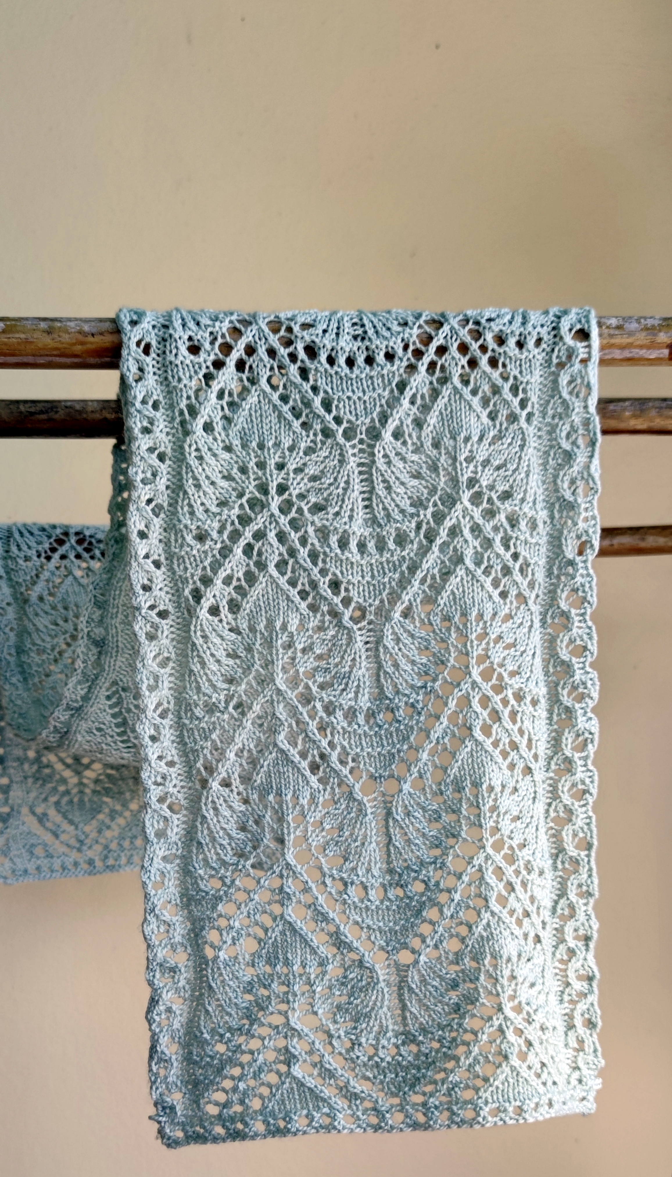 A lacy leaf scarf in a pale silver is draped over a wooden rod.