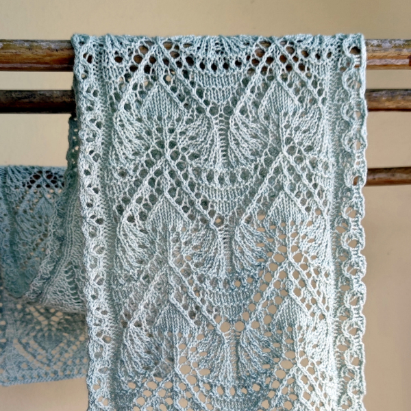 A lacy leaf scarf in a pale silver is draped over a wooden rod.