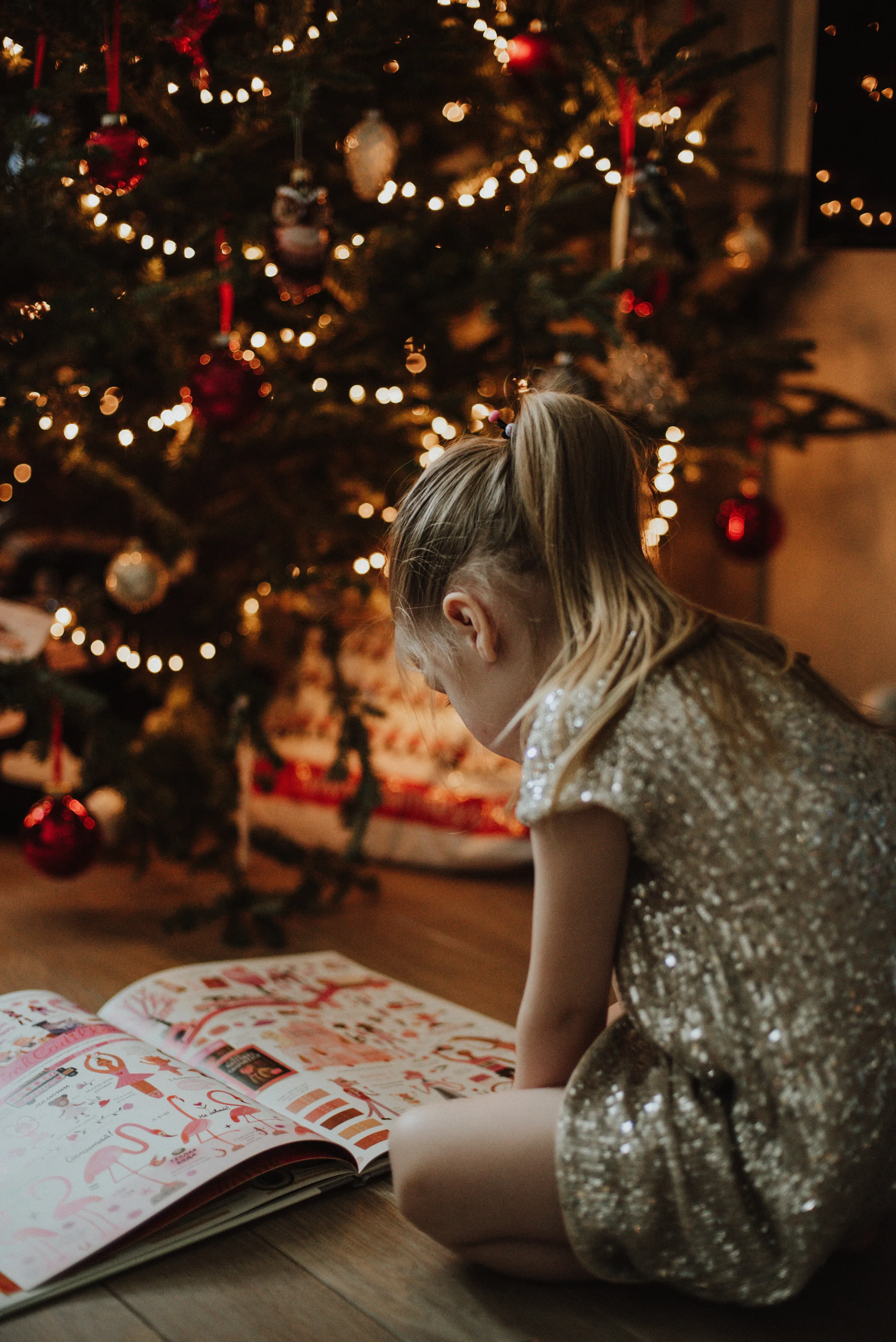 A girl in a shiny silver dress reads a book on the floor in front of a lit Christmas tree.