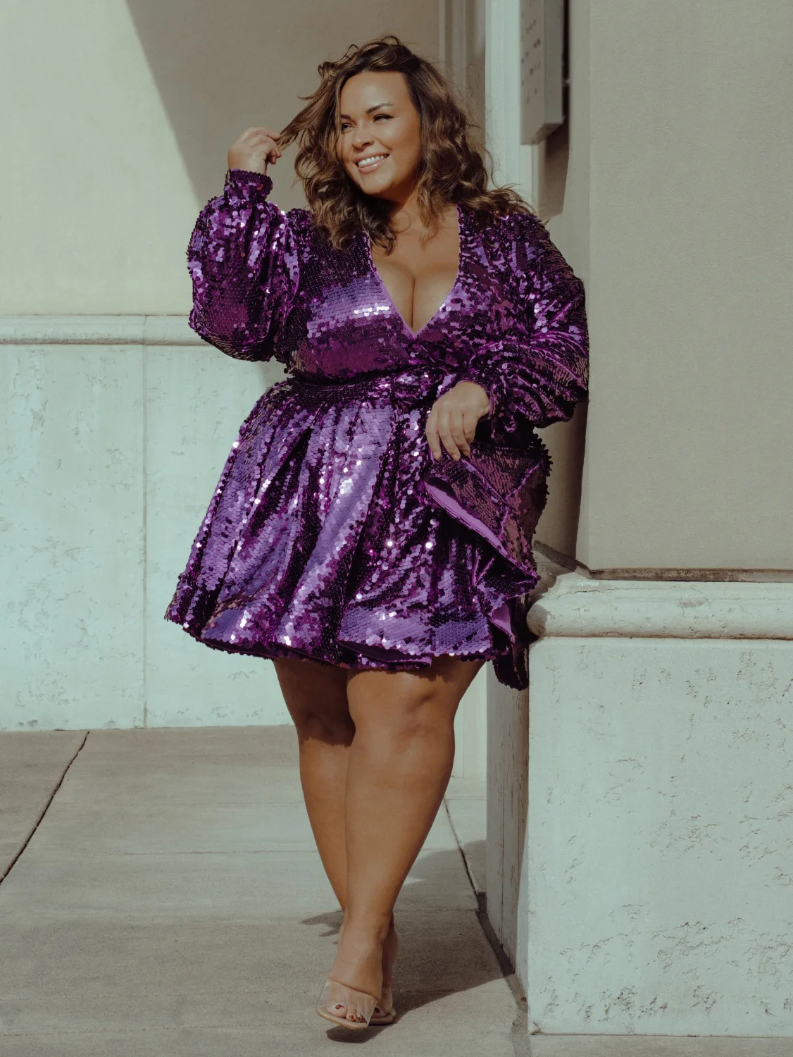 A woman leans against a wall, wearing a short purple sparkly dress.