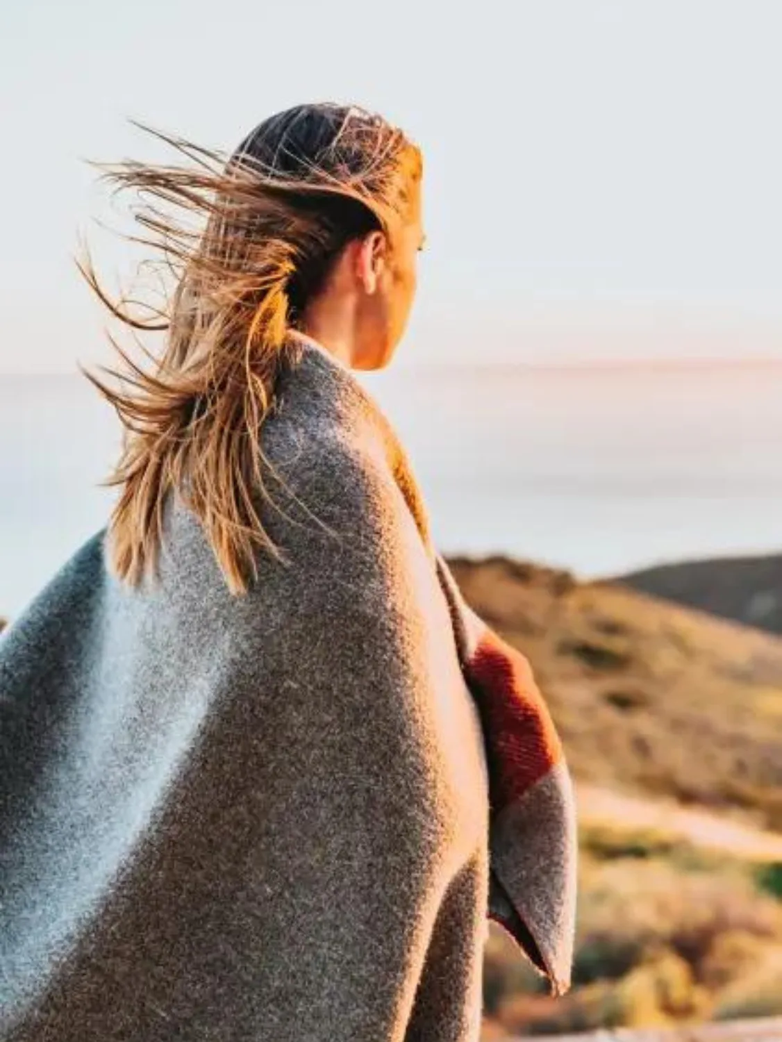 A woman stares off into the sandy distance wearing a grey wool blanket wrapped around her shoulders. Her hair is windblown.